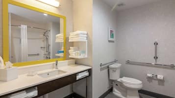 A modern bathroom with a large mirror, white sink, and toilet. There are towels and toiletries on the counter, and a shower area is visible.