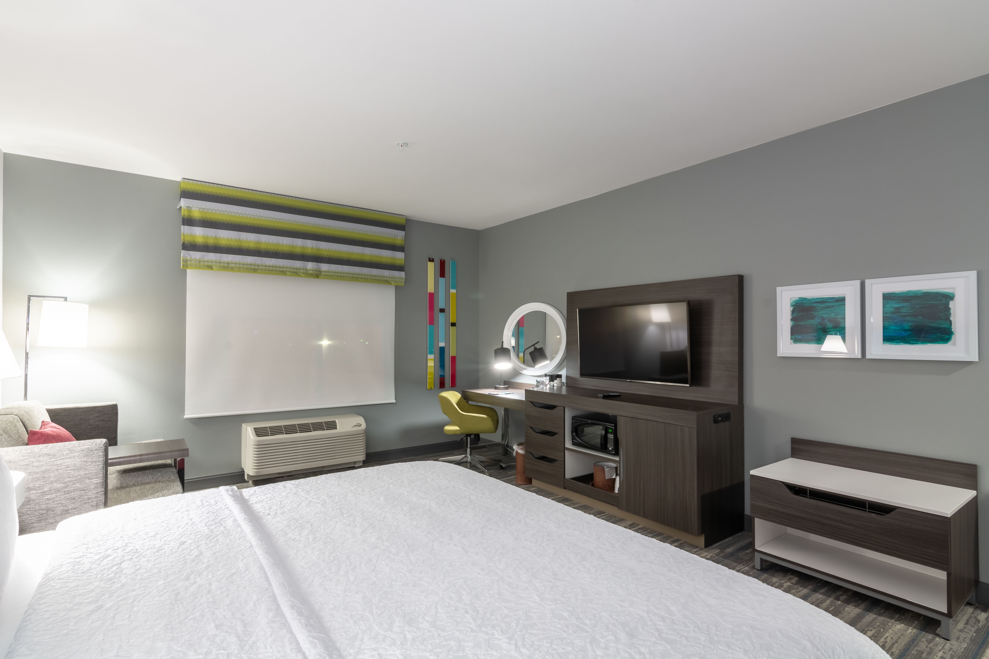 Une chambre d’hôtel moderne, dotée d’un grand lit, d’un bureau avec une chaise, d’une télévision, d’un miroir et d’œuvres d’art accrochées aux murs.