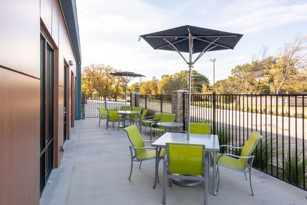 Une terrasse extérieure avec des tables et des chaises, un espace salon couvert et une vue sur un parking et des arbres.