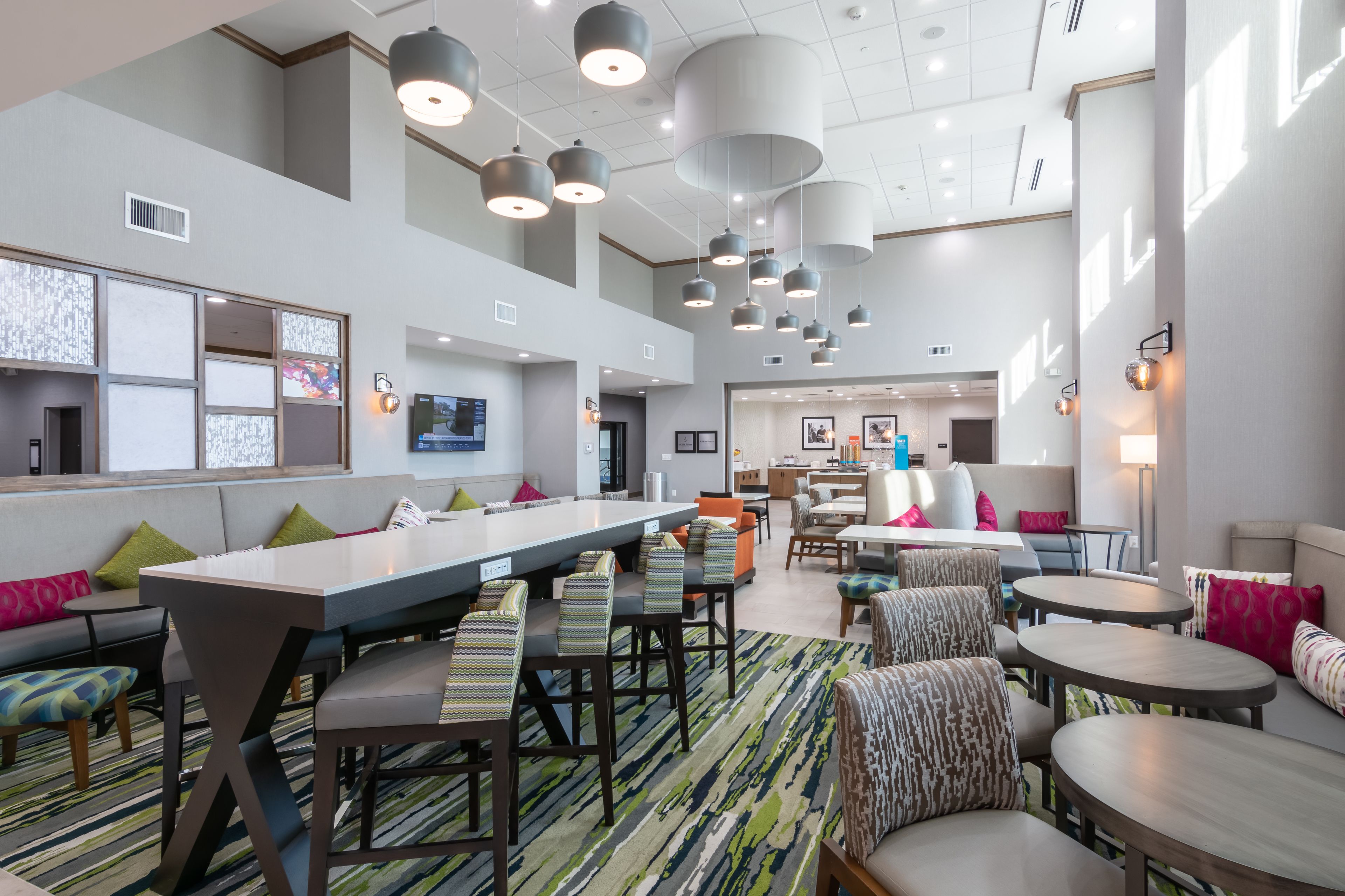 A modern hotel lobby with a long reception desk, multiple seating areas, and a high ceiling with hanging lights.