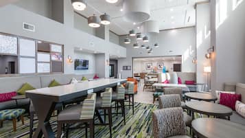 A modern hotel lobby with a long reception desk, multiple seating areas, and a high ceiling with hanging lights.