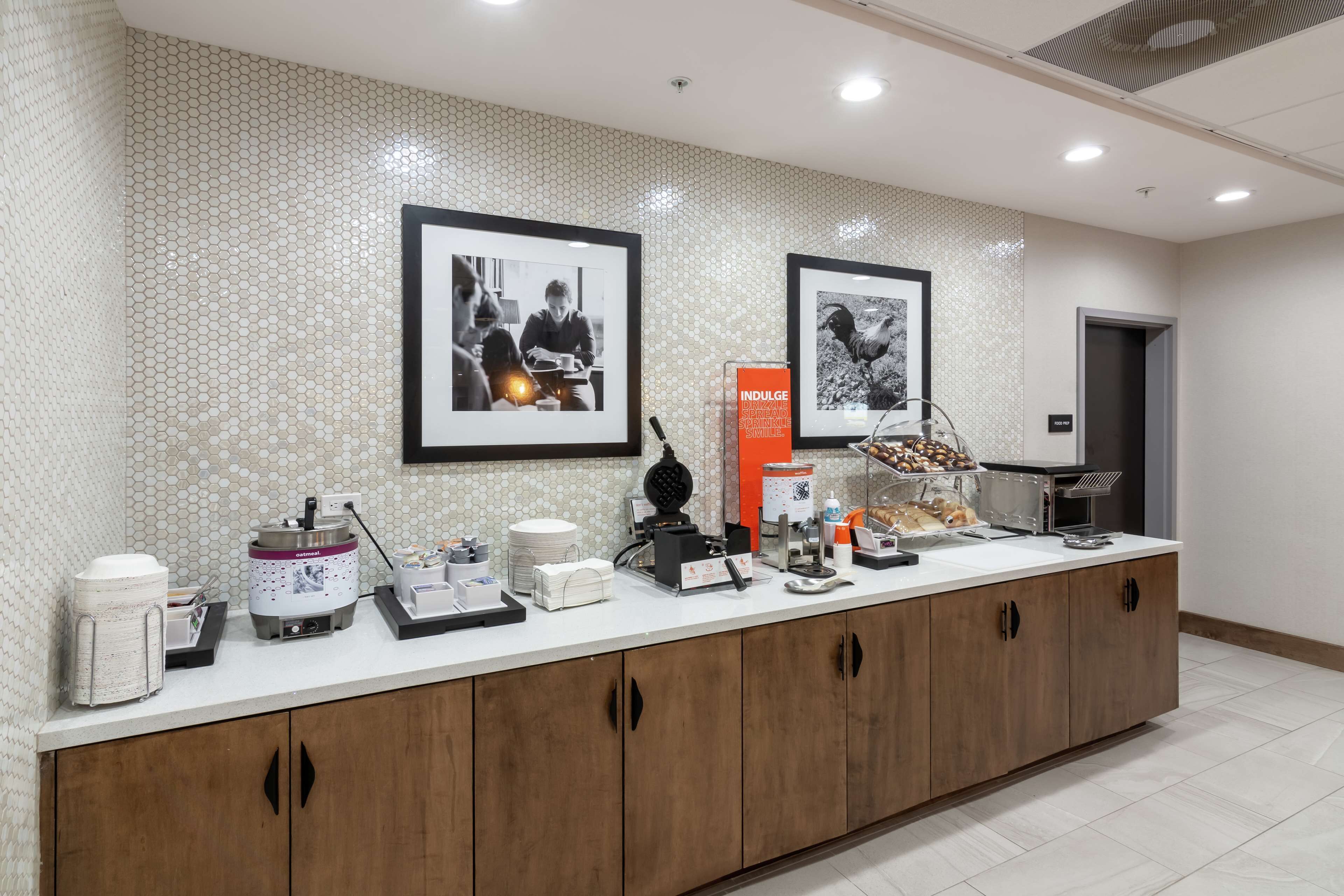 Un espace d’accueil d’hôtel comprenant un comptoir, des machines à café, un présentoir de fruits et des tableaux encadrés accrochés au mur.