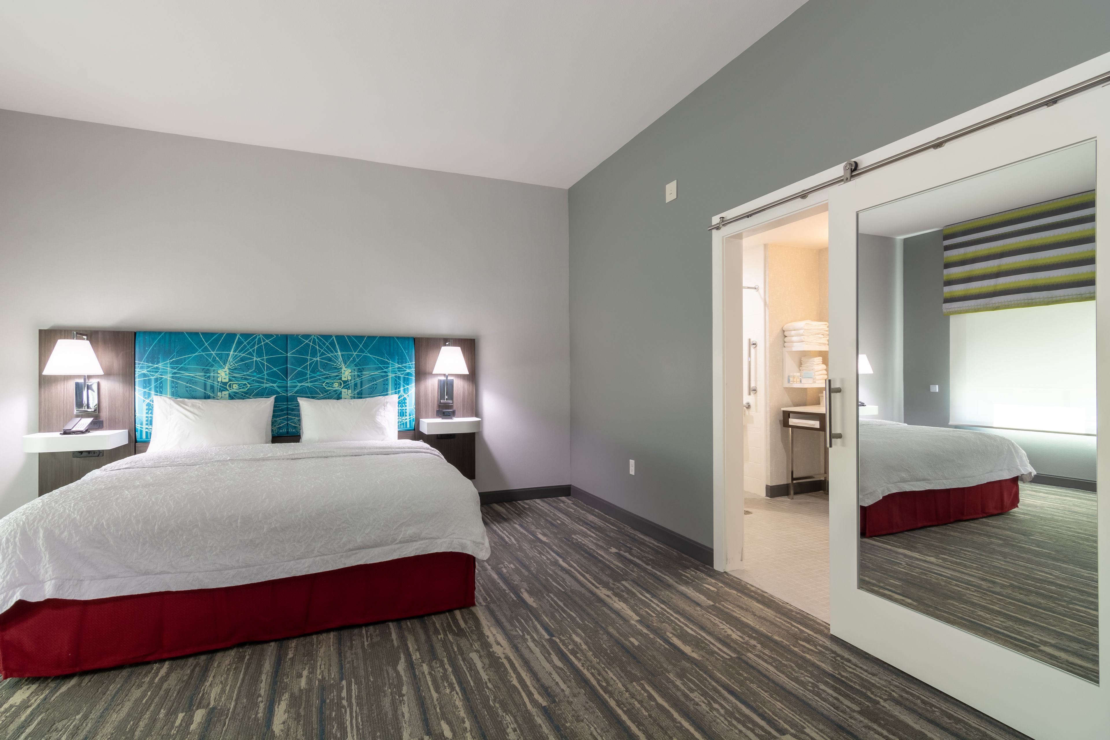 Una camera d'albergo con un letto grande, una parete azzurra, due lampade sui comodini, uno specchio e un ingresso che porta al bagno.