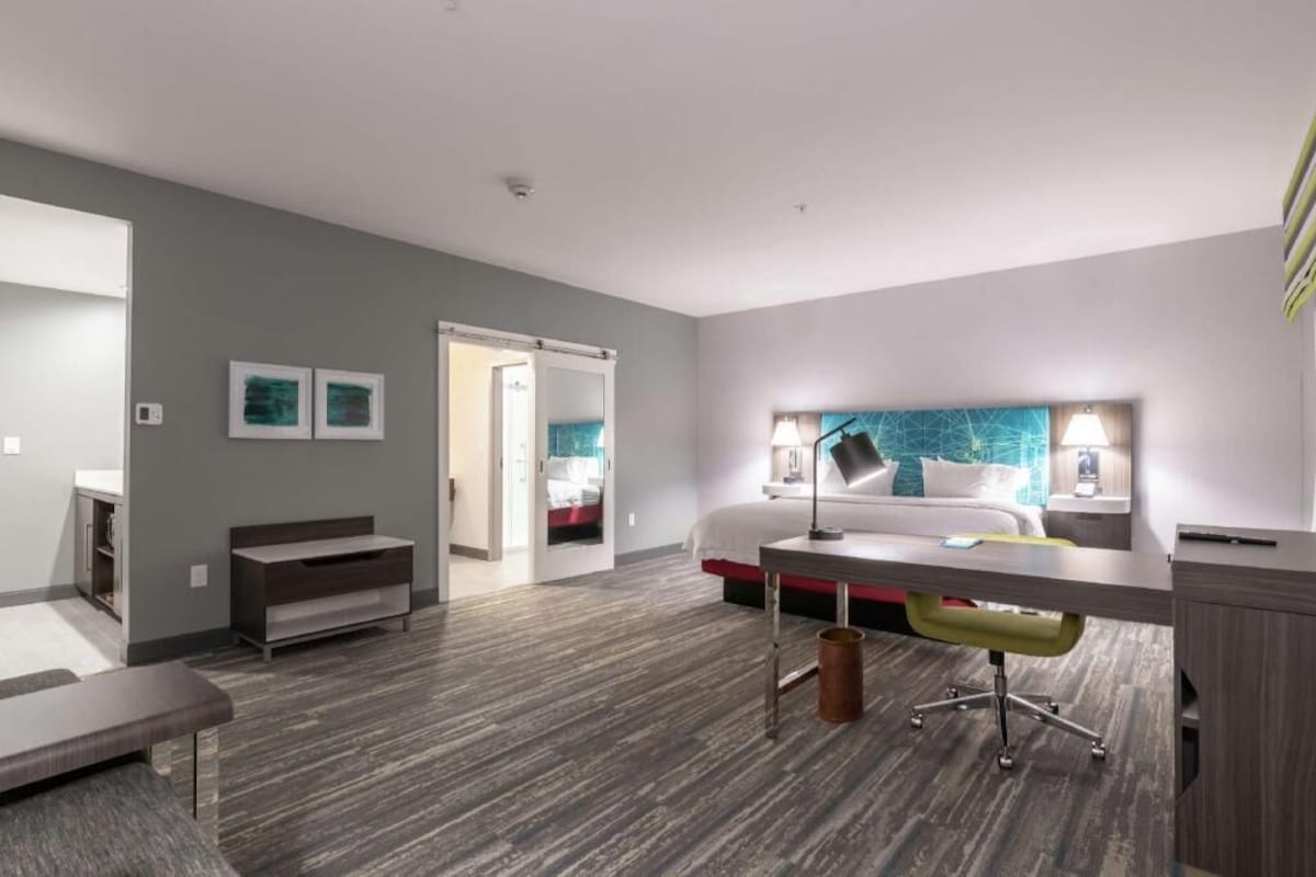 A modern hotel room with a bed, desk, chair, and a large window.