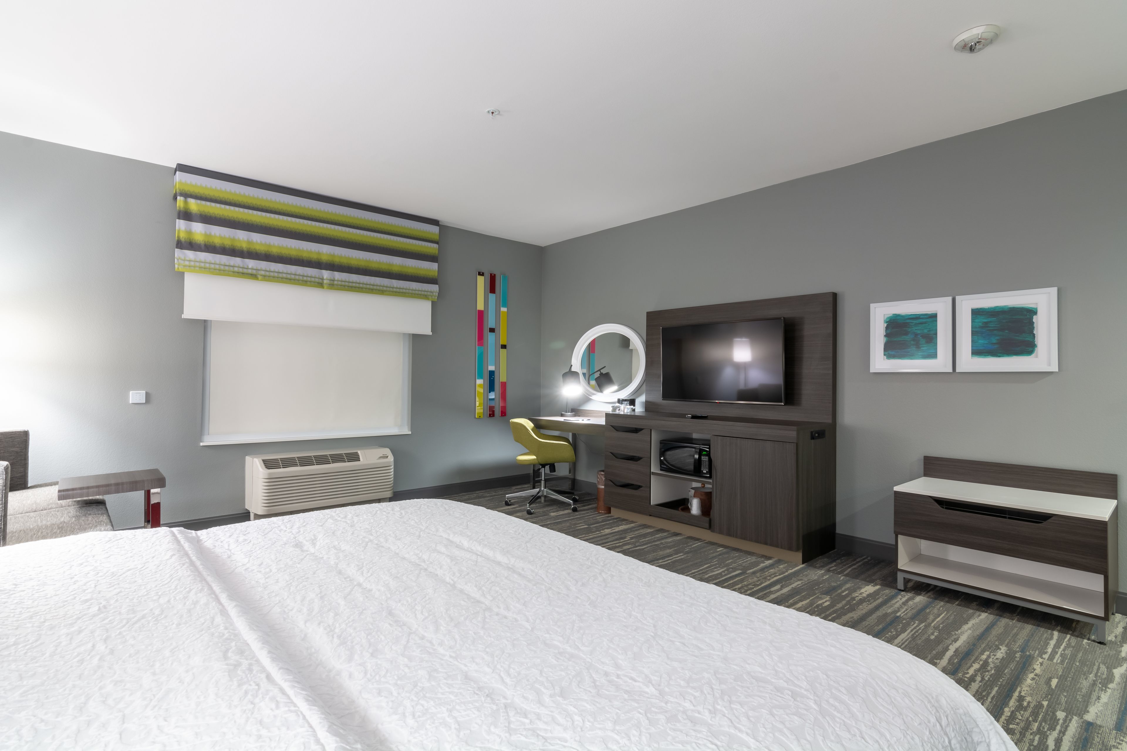 A modern hotel room with a large bed, a desk with a chair, a television, a mirror, and artwork on the walls.