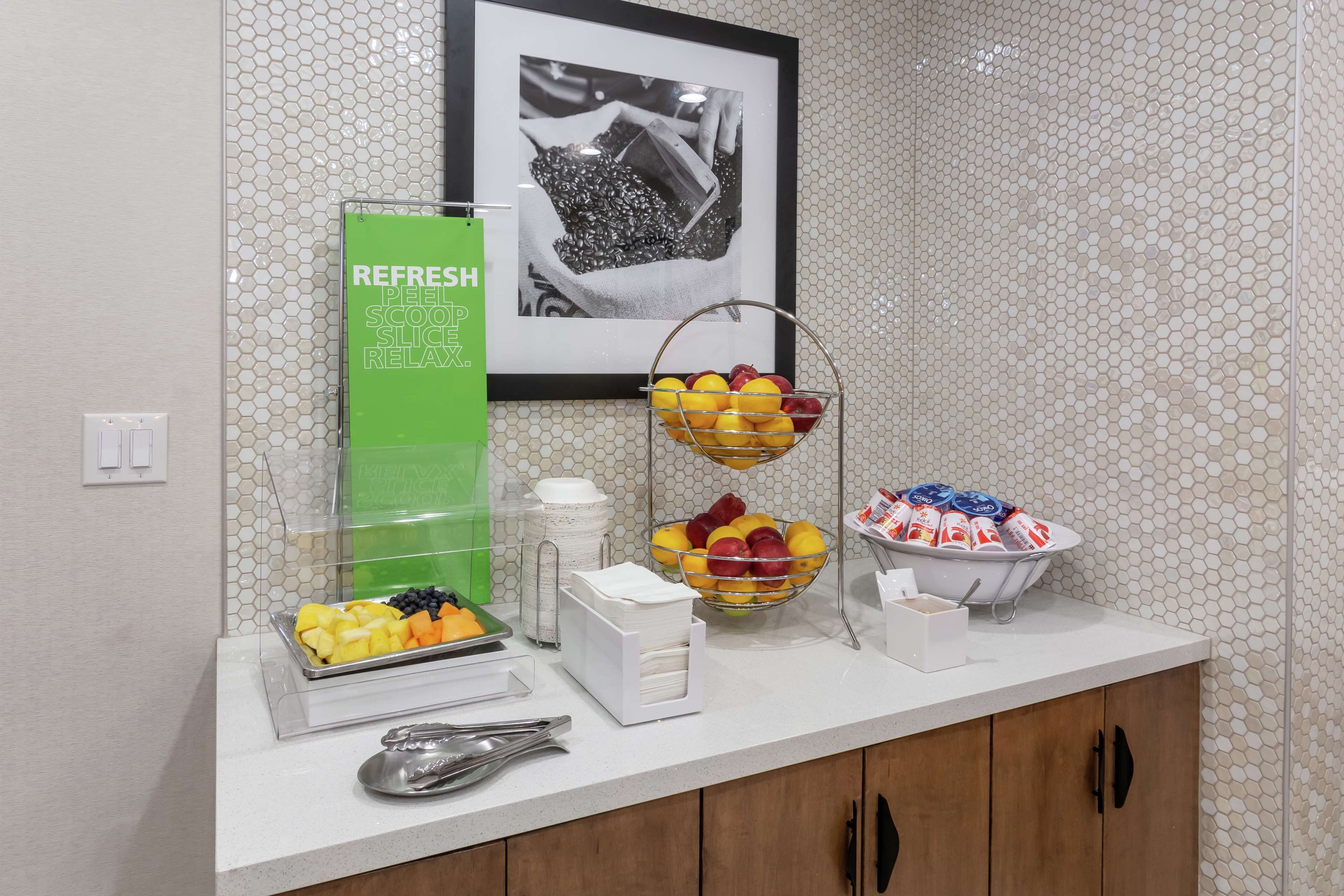 Un buffet per la colazione con vassoi di frutta, un erogatore di bevande fresche verdi e un quadro appeso al muro.