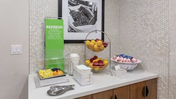 Un buffet per la colazione con vassoi di frutta, un erogatore di bevande fresche verdi e un quadro appeso al muro.