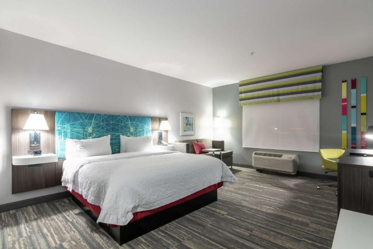 A modern hotel room with a large bed, bedside tables, a desk, a chair, a window with blinds, and a wall-mounted air conditioner.