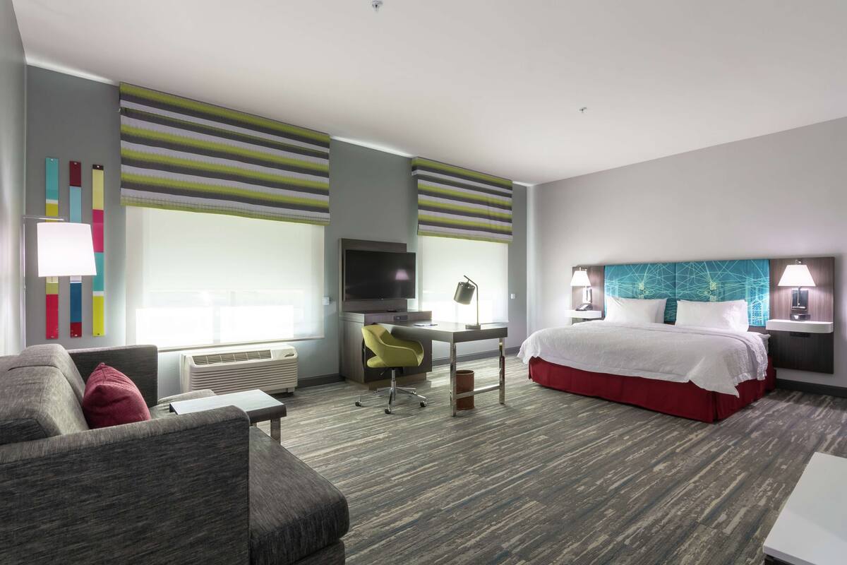 A modern hotel room with a grey sofa, a desk, a bed with a red skirt, and a TV.