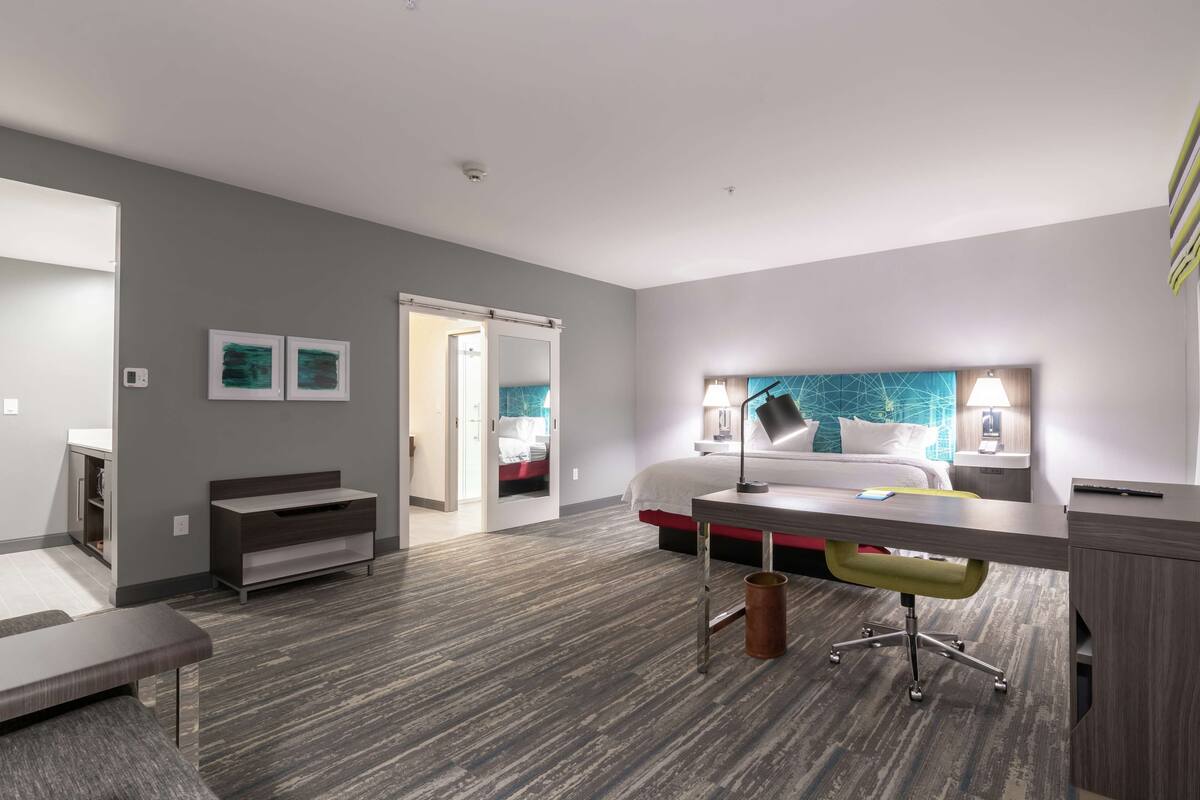 A modern hotel room with a bed, desk, and chair. There is a door leading to another room, and a small seating area with a bench.