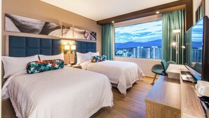 A hotel room with two beds, a desk, a chair, a TV, and a large window with a city view.