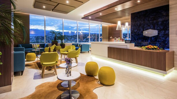 A modern hotel lobby with a reception desk, seating area with yellow chairs, and large windows offering a city view.