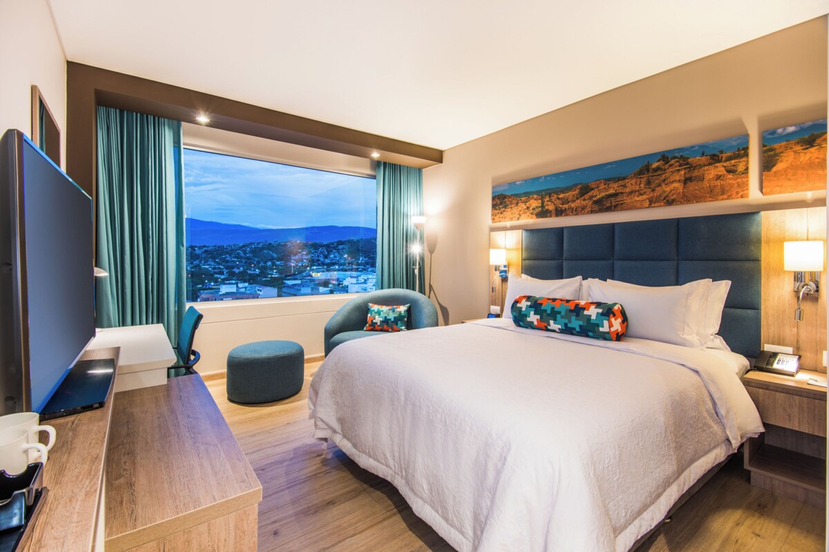 Ein Hotelzimmer mit einem großen Bett, einem Schreibtisch mit einem Laptop, einem Sessel und Blick auf eine Stadtlandschaft.