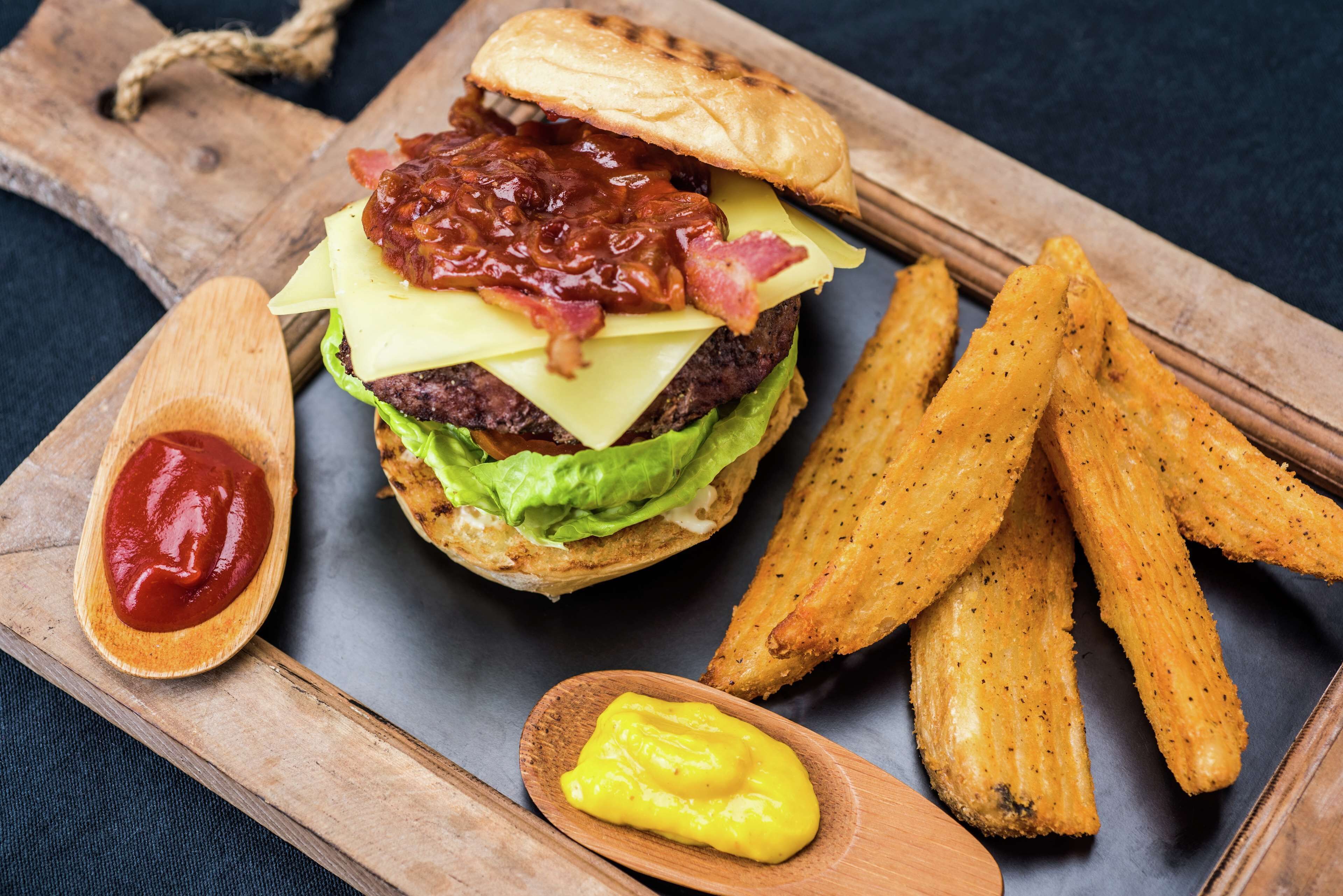 Een hamburger met bacon, kaas en sla, geserveerd met friet en sauzen op een houten plank.