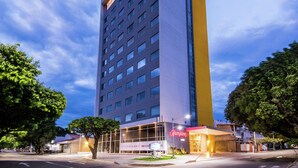 A tall hotel building with a prominent 'Hampton' sign, featuring a modern design with a mix of glass and solid facade, and a visible entrance with a well-lit area.