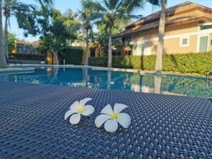 Zona de piscina con dos flores blancas sobre una superficie tejida, rodeada de palmeras y un edificio al fondo.