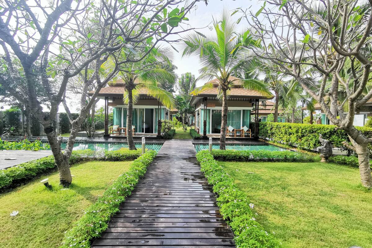 Un camino que lleva a dos villas con piscina, rodeado de exuberante vegetación y palmeras.