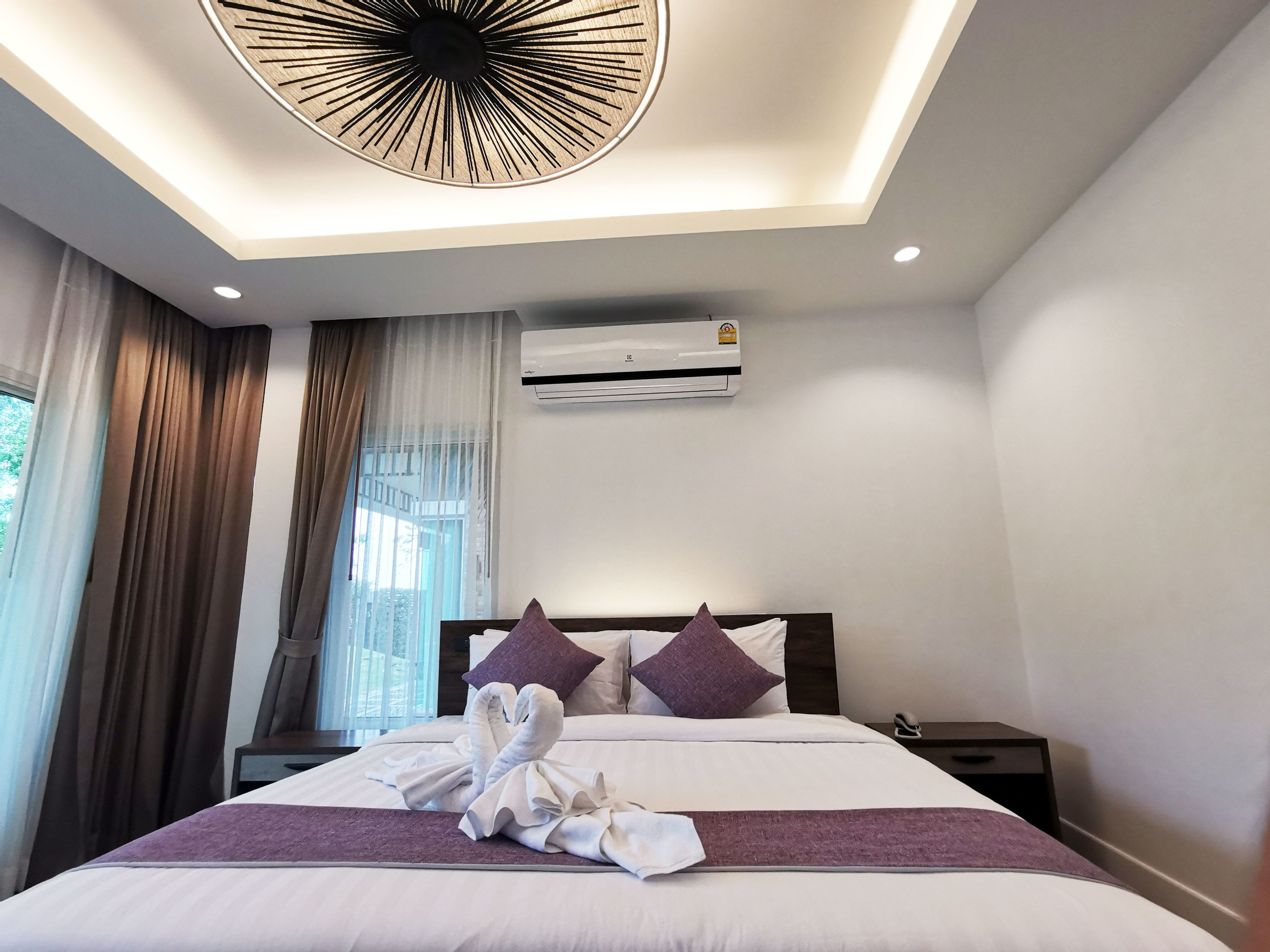 A hotel room with a bed, a ceiling light fixture, an air conditioning unit, and a window with curtains.