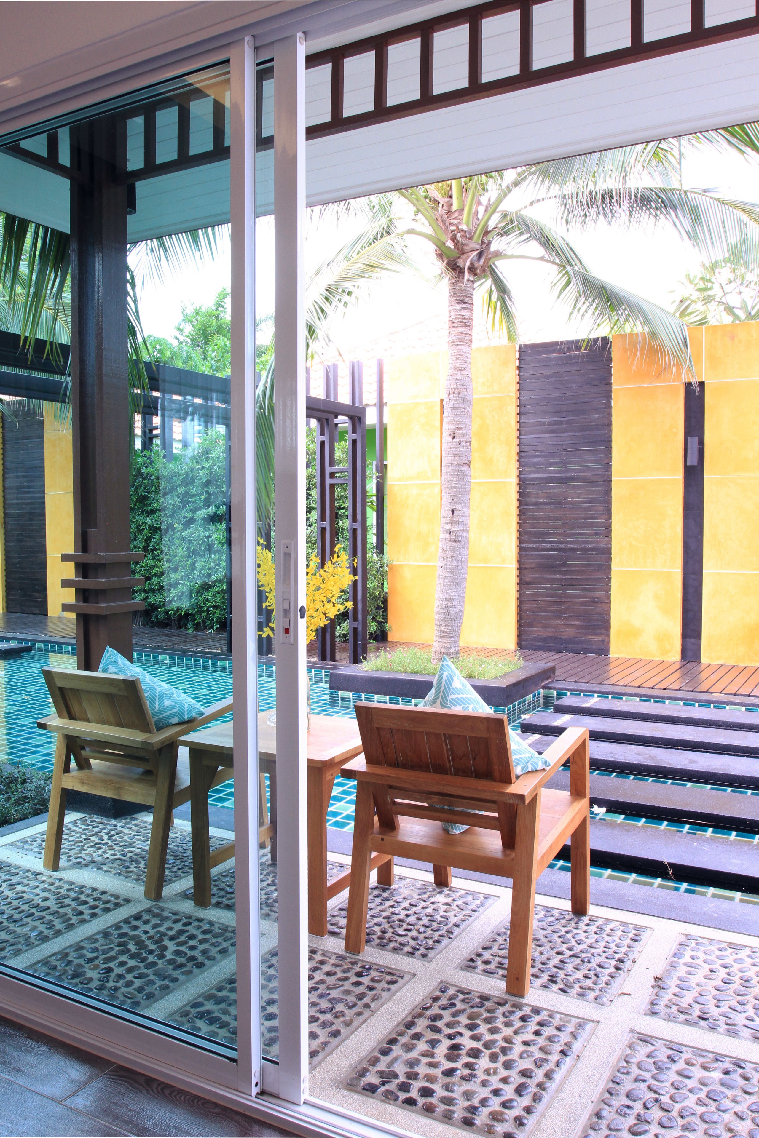 A glass-enclosed patio with two wooden chairs, a small table, and a pool area with a yellow wall and palm trees in the background.