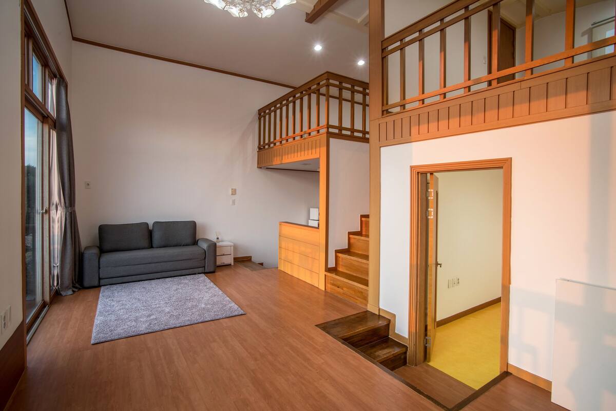 Amplia sala de estar con un sofá gris, piso de madera, una escalera y vista a través de un ventanal.