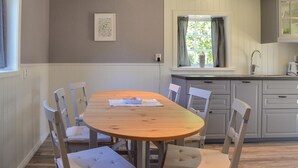 A dining area with a wooden table and white cushioned chairs, a window with curtains, and a kitchen with grey cabinets. - Cozy home in Herräng with WiFi (Herräng)