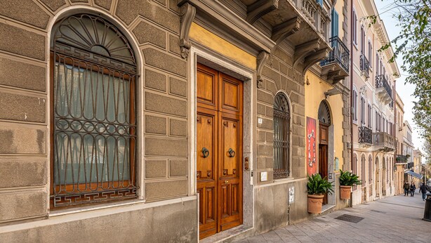 Fachada de un edificio histórico con una puerta de madera grande, una ventana con rejas decorativas y una planta en maceta en la acera.