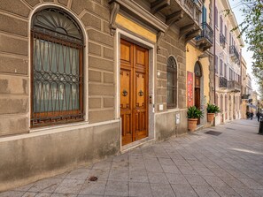 Fachada de um edifício histórico com uma grande porta de madeira, uma janela com grades de ferro decorativas e um vaso de planta na calçada.