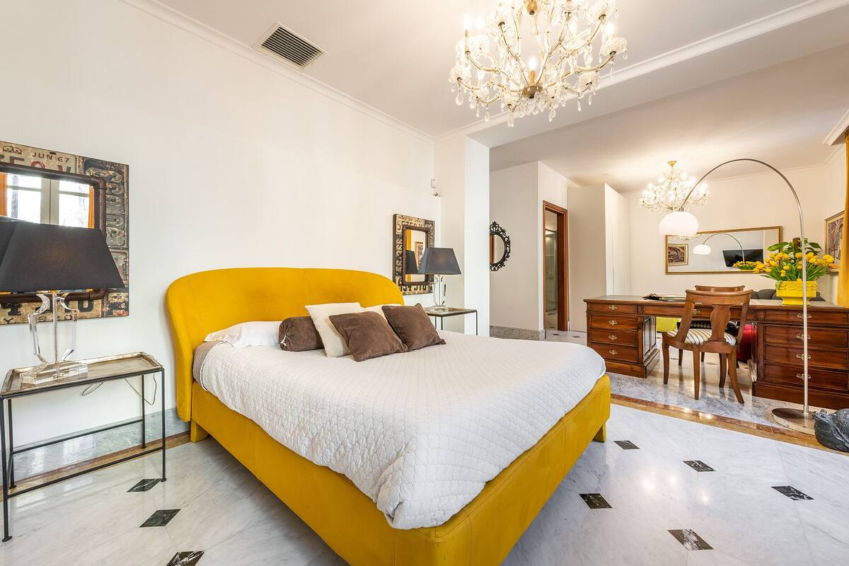A hotel room with a large bed, a desk, a chair, a chandelier, and a mirror.