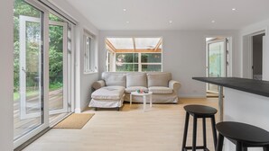 A modern living room with a sofa, ottoman, and a small table. There is a large window providing natural light and a view of the outdoors.