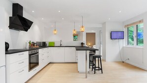 A modern kitchen with white cabinets, a black range hood, and a wooden floor. There are two pendant lights, a TV mounted on the wall, and a window with a view of greenery.