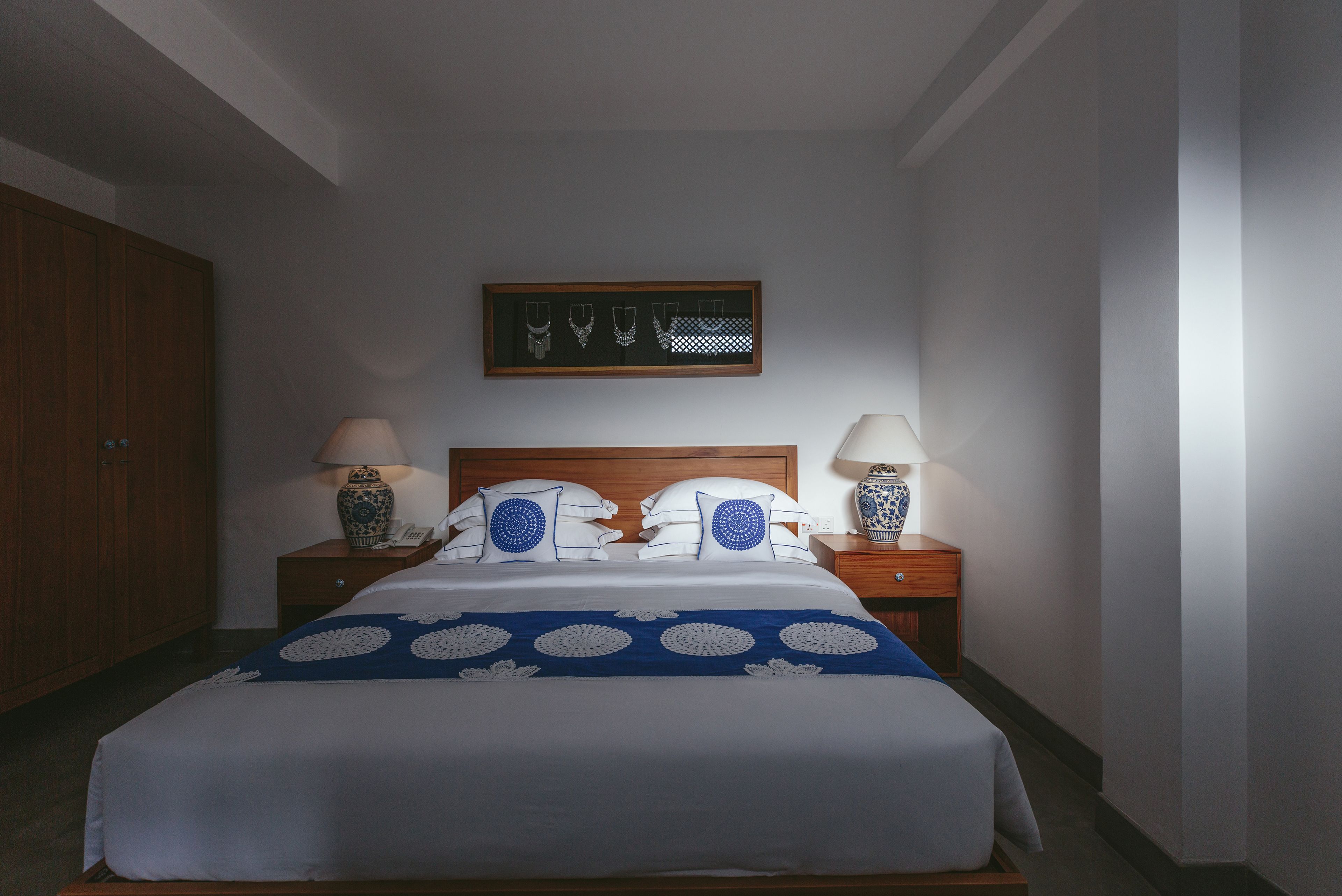 A neatly made bed with a blue and white patterned bedspread, two bedside lamps, a framed picture above the bed, and a wooden wardrobe to the left.