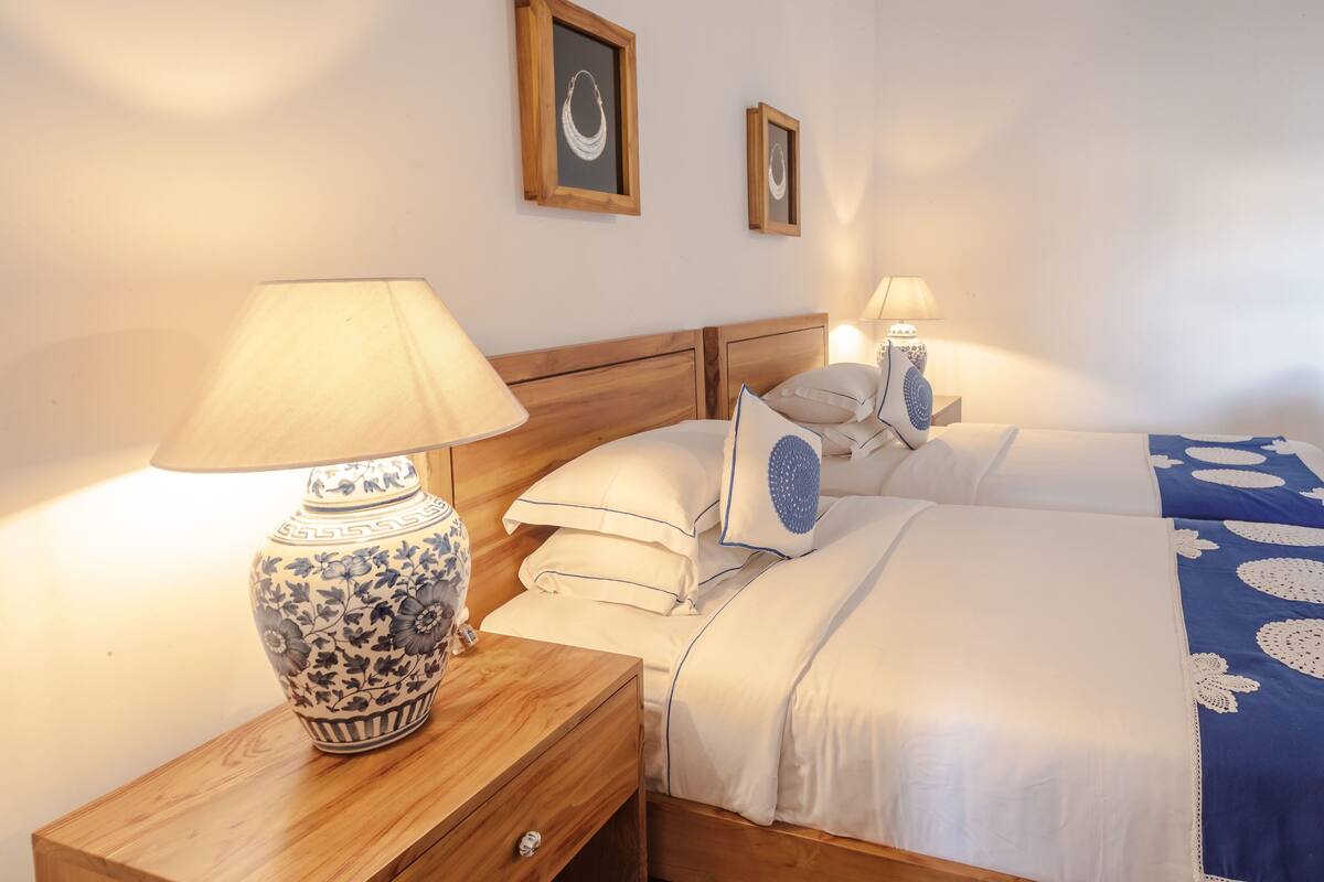 Un lit bien fait, avec une literie blanche et bleue, une table de chevet en bois surmontée d’une lampe, et deux tableaux encadrés accrochés au mur.