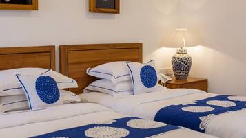A hotel room with two beds, each with white bedding and blue decorative pillows. There is a wooden headboard, a bedside table with a lamp, and a framed picture on the wall.