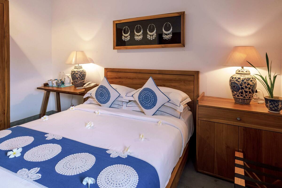 Un lit bien fait, recouvert d’une couvre-lit à motifs bleus et blancs, avec deux oreillers assortis et une tête de lit en bois. Une petite table avec une lampe et un vase se trouve sur le côté droit du lit.