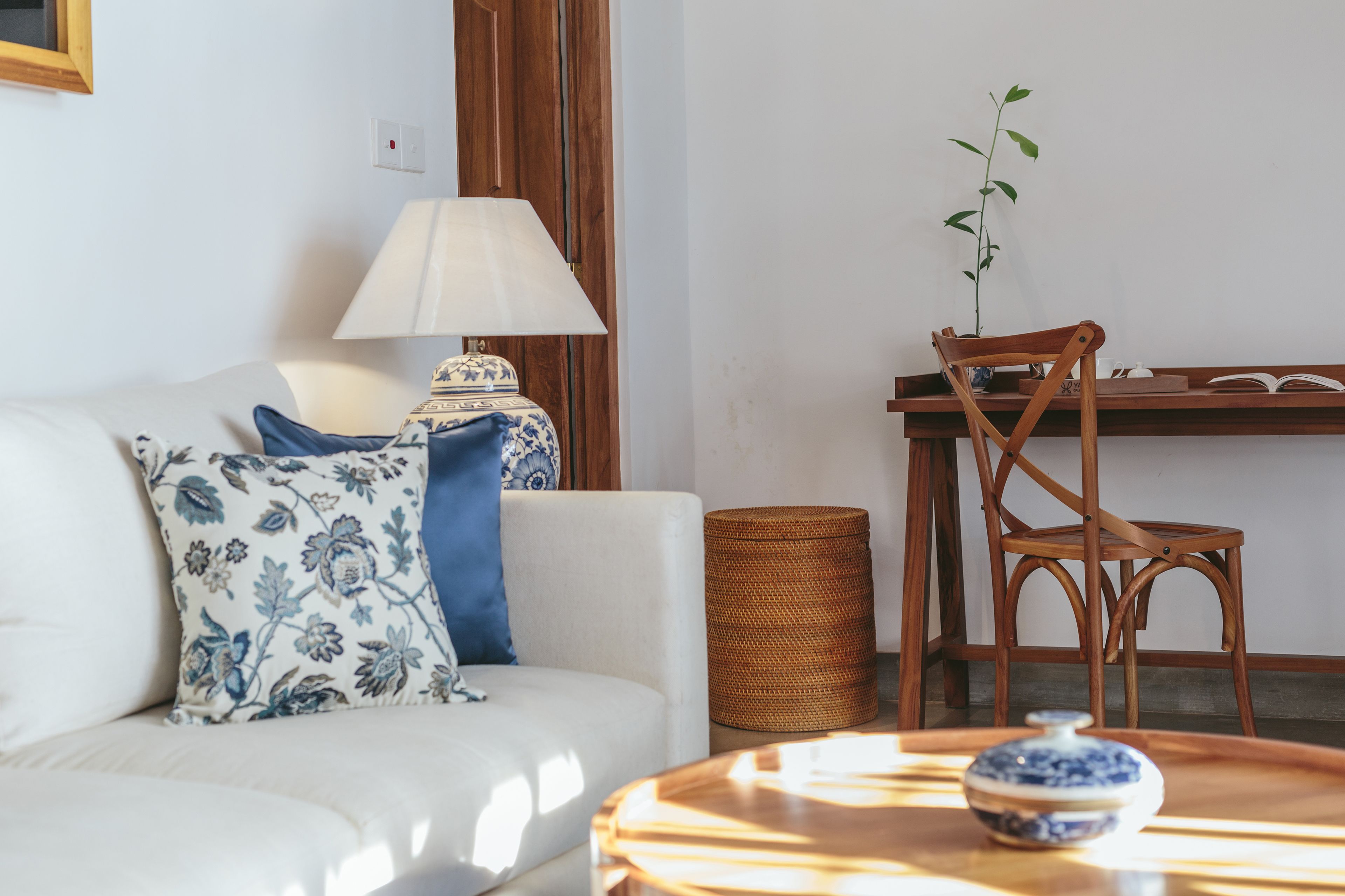 A living room with a white sofa, a blue and white patterned pillow, a wooden chair, a small table with a blue and white vase, and a lamp.