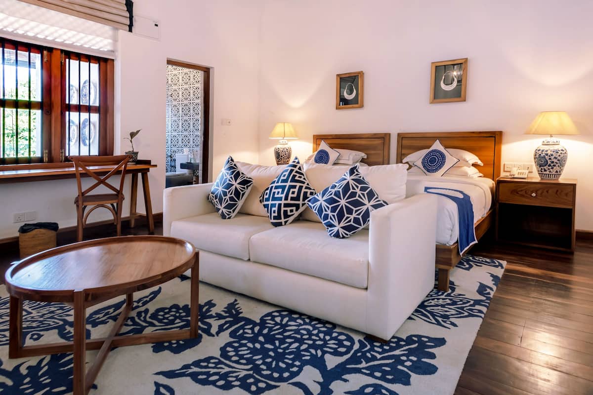 Une chambre d’hôtel avec un canapé blanc, un tapis à motifs, une table d’appoint en bois et un lit agrémenté de coussins décoratifs.