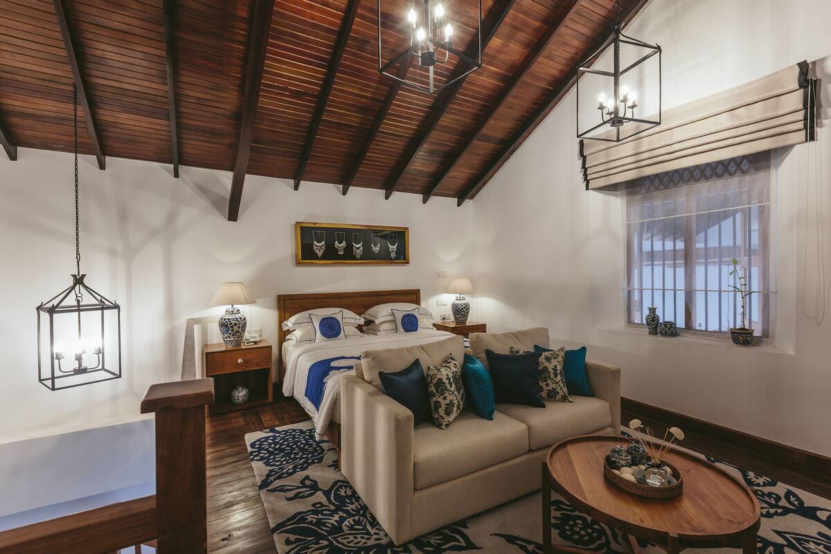 Une chambre confortable avec un plafond en bois, un lit recouvert d’une literie bleue et blanche, un canapé beige et une petite table ronde agrémentée d’objets décoratifs.