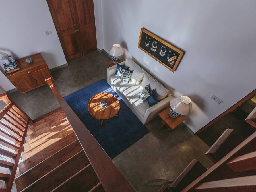 A modern living room with a wooden staircase, a blue rug, a white sofa with patterned cushions, and a wooden side table.