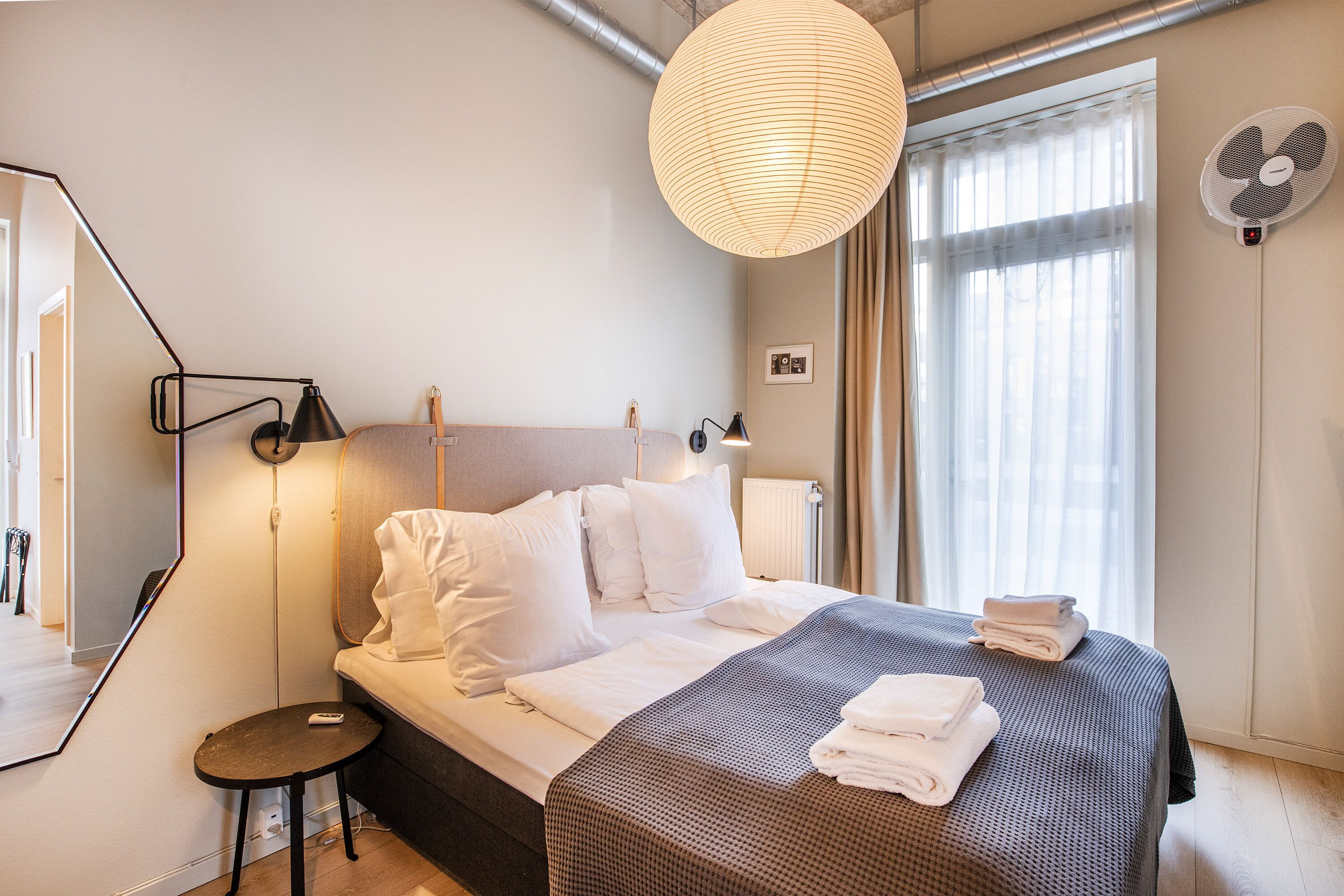 A neatly made bed with white linens, a bedside table with a lamp, and a large window with curtains.