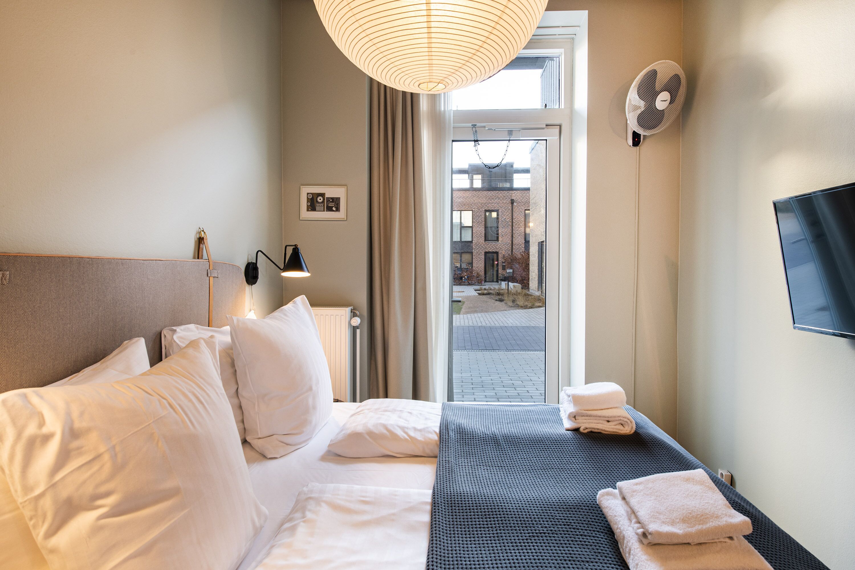 Et hotelværelse med en seng, puder, en sengetavlelampe, en ventilator, et fjernsyn og udsigt ud.