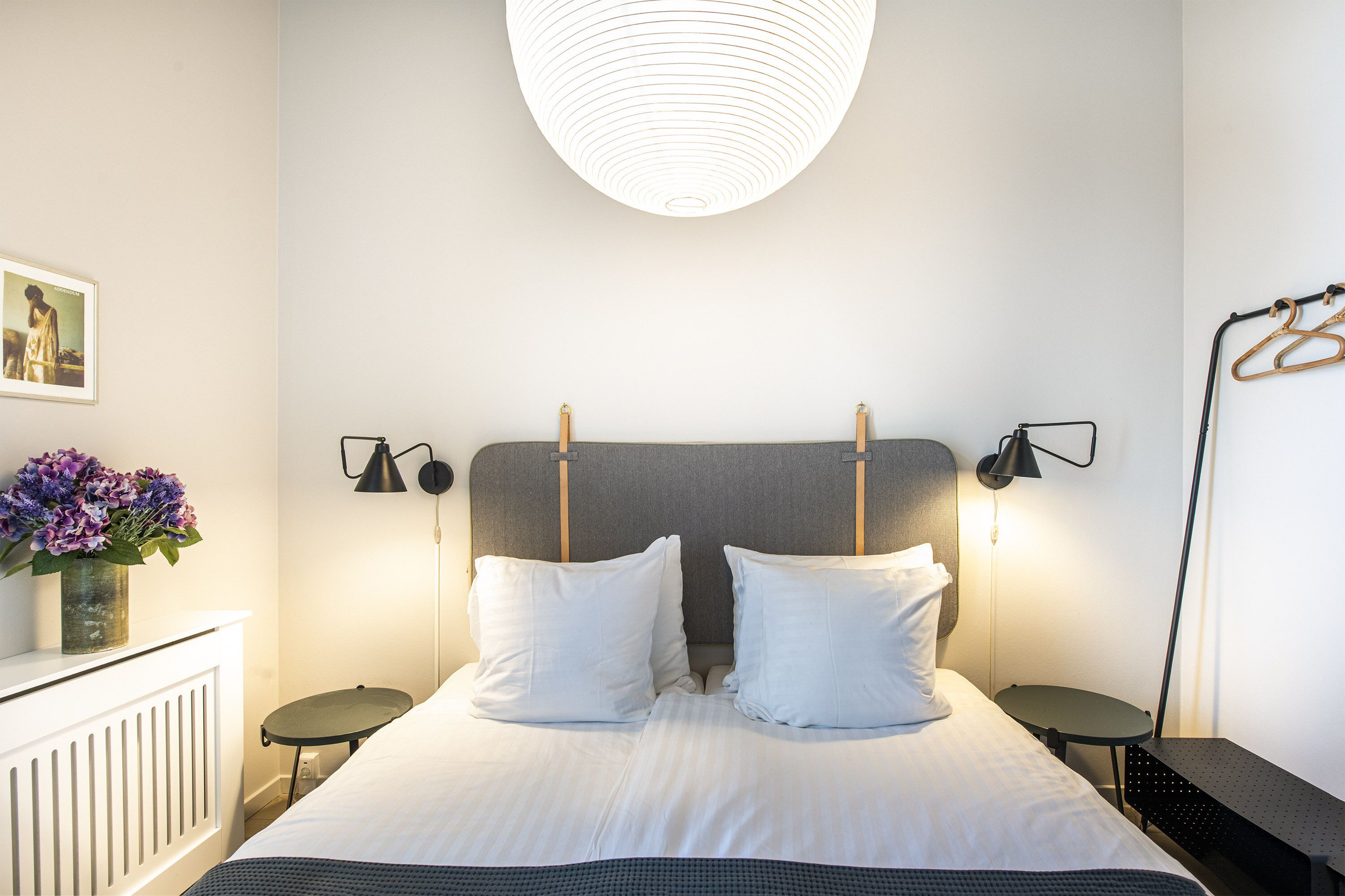 Una cama bien hecha con ropa de cama blanca, dos mesitas de noche, una lámpara de pared y un perchero.