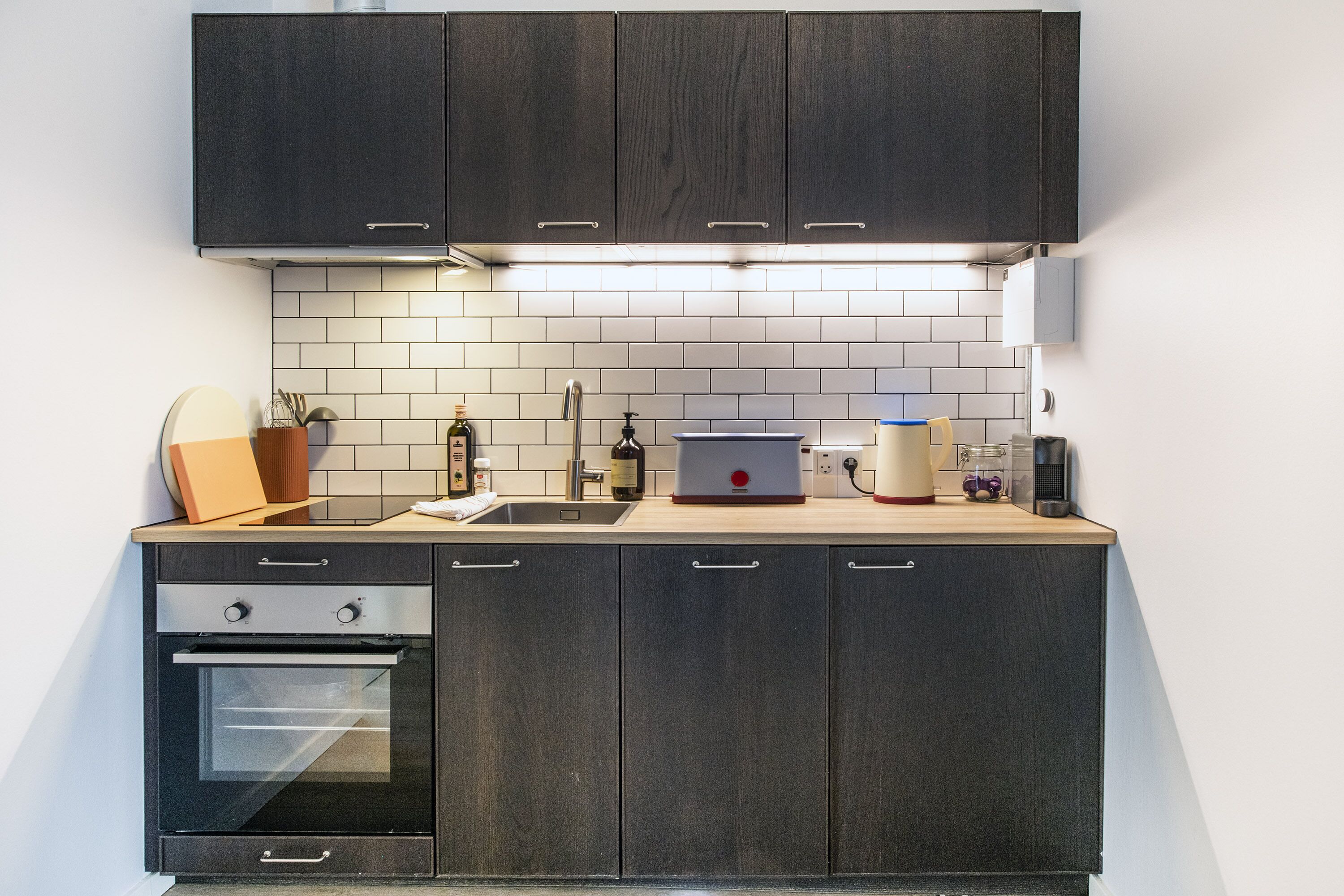 Una cocina moderna con gabinetes de madera oscura, horno empotrado, pileta y una encimera con diversos electrodomésticos y utensilios.