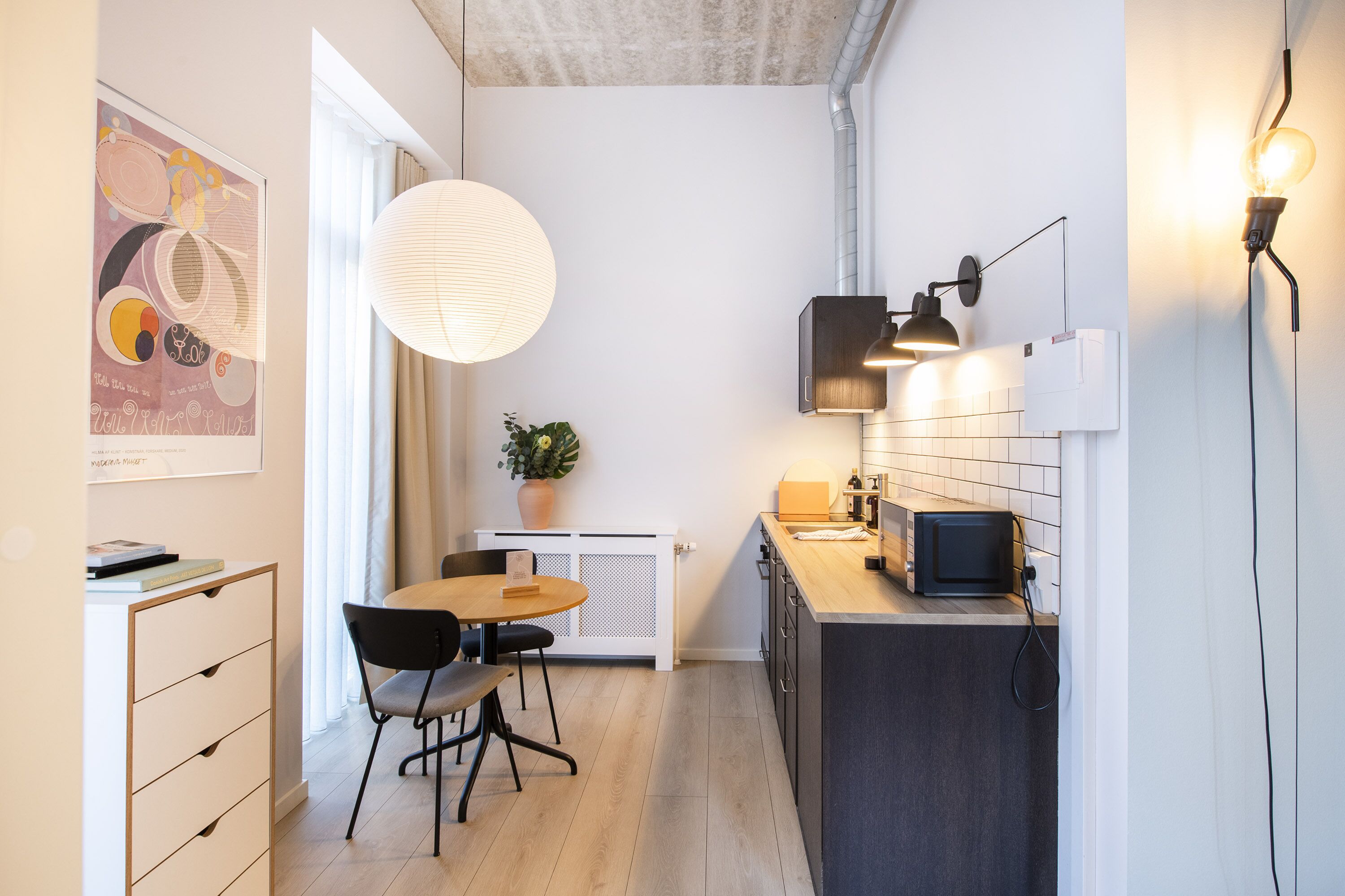 A compact kitchen with a dining area, a white radiator, a potted plant, and a modern pendant light.