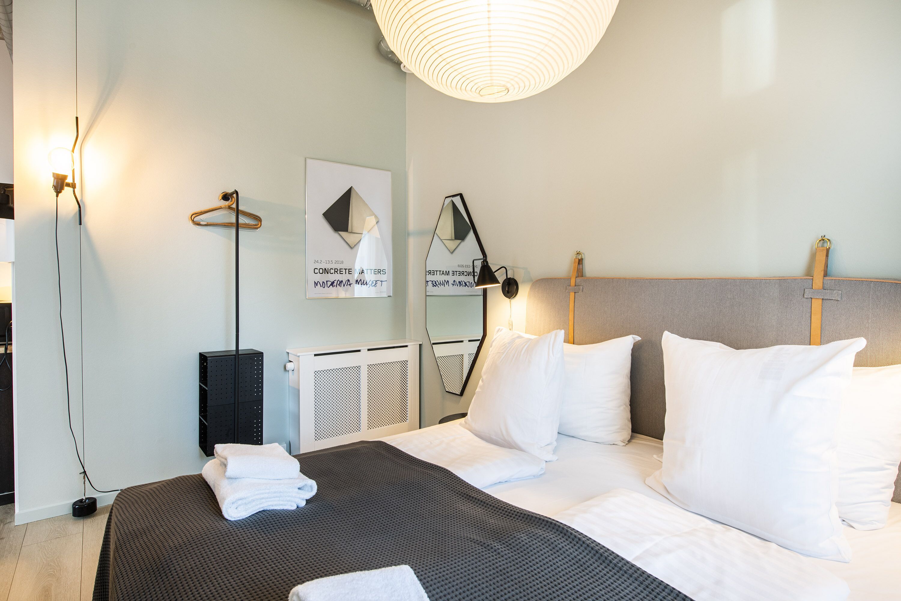 Quarto de hotel moderno com cama bem arrumada, luminária de cabeceira, cabideiro e uma obra de arte emoldurada na parede.