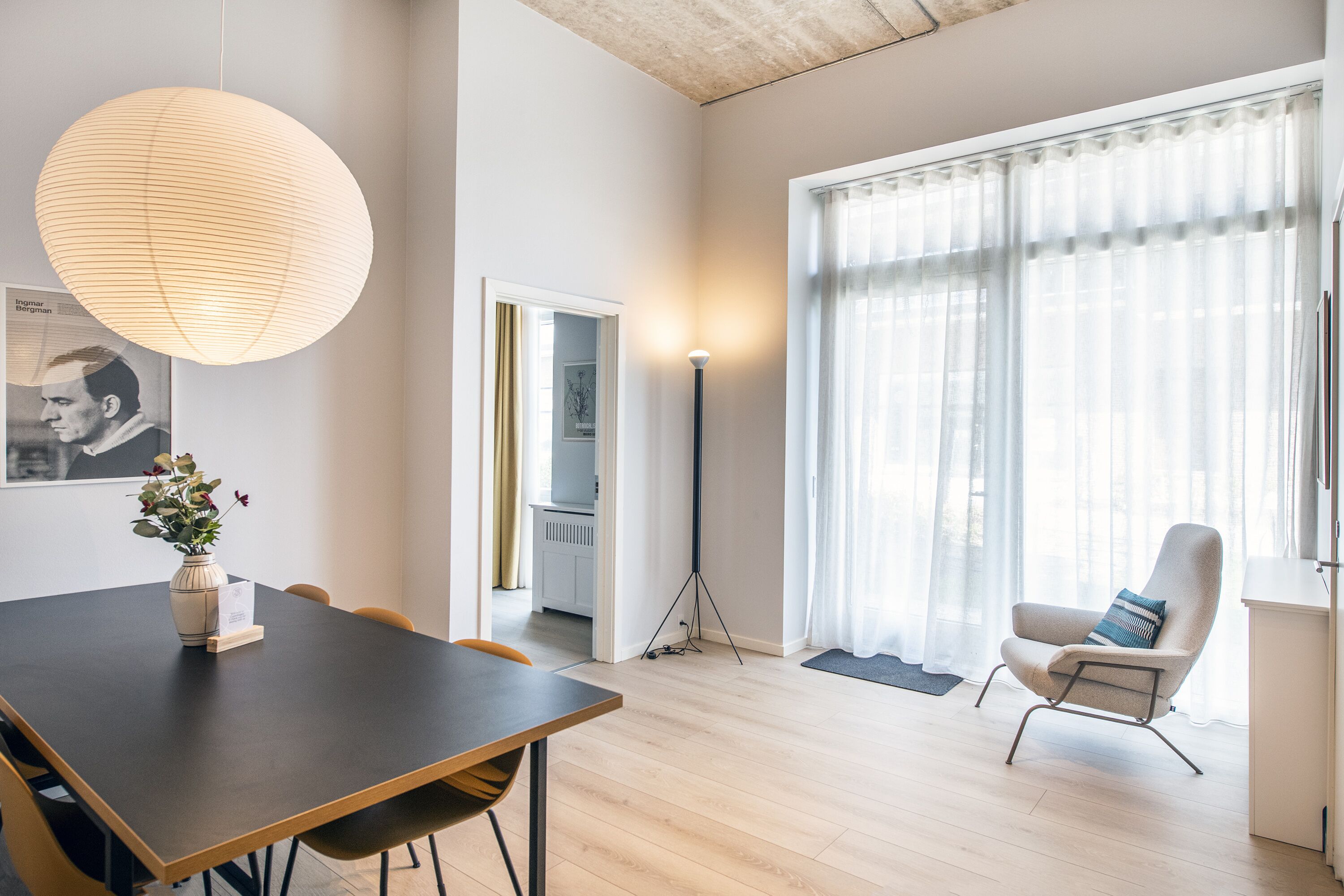 Un salon moderne avec une table à manger en bois foncé, une grande lanterne ronde en papier, un lampadaire, un fauteuil confortable et une grande fenêtre ornée de rideaux légers.