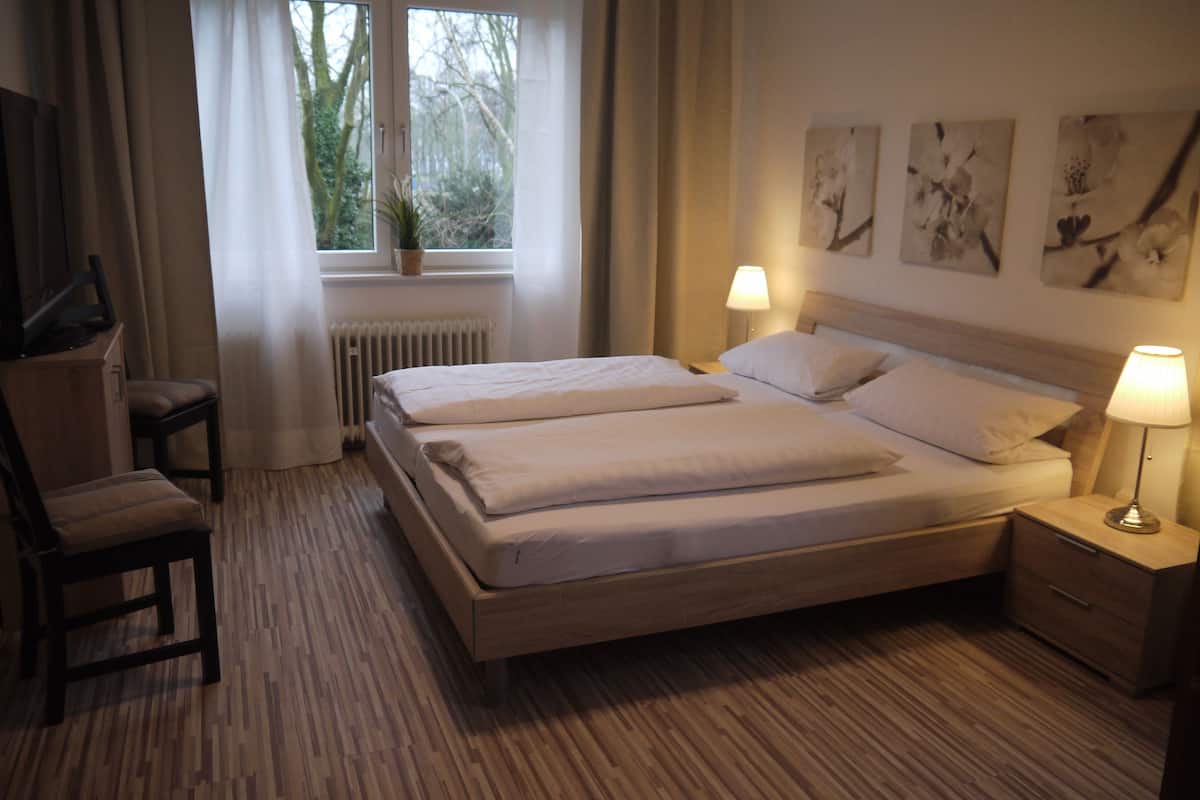 A hotel room with a double bed, two bedside tables with lamps, a chair, a window with curtains, and a radiator.