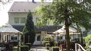 Een gebouw met een betegeld dak, een overdekt terras met tafels en stoelen, en een pad naar de ingang.