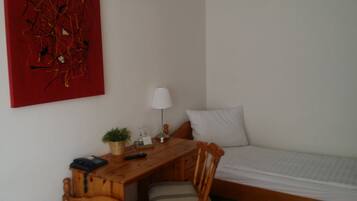 Un dormitorio pequeño y ordenado con un escritorio de madera, una silla, una mesita de noche con lámpara, una cama y un cuadro abstracto rojo en la pared.
