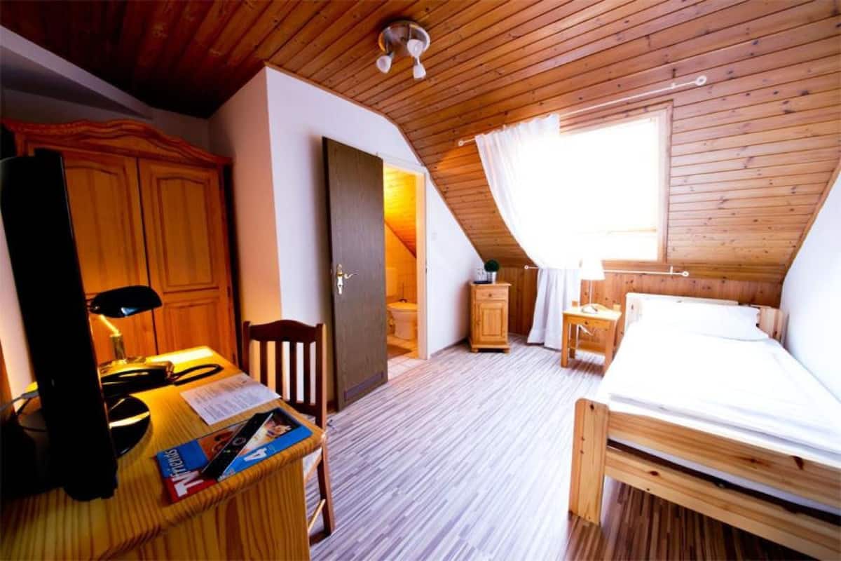 A bedroom with a wooden ceiling, a bed, a desk with a chair, a wardrobe, and a window.