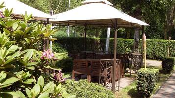 Un espace extérieur abrité, aménagé avec des tables et des chaises, et entouré de verdure et de haies.
